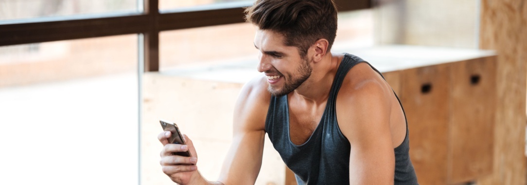 A photo of a smiling man relaxing during a workout near the weights station with a mobile phone in his hand
