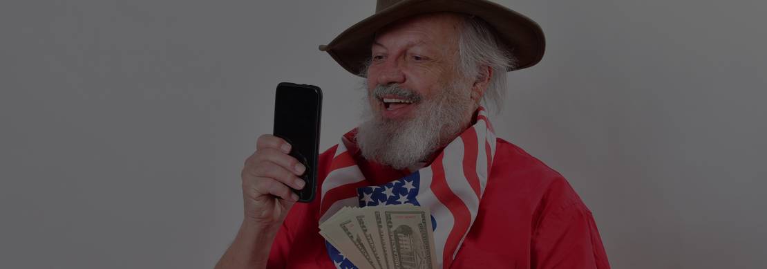old cowboy playing Juicy Stakes Poker on his phone and holding cash he's won in one hand
