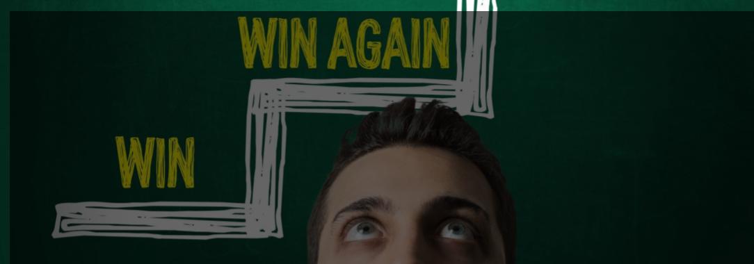 A photo of a man looking up with ‘Win’, ‘Win Again’, ‘Win More’ in a stepped format on green board on the wall behind him