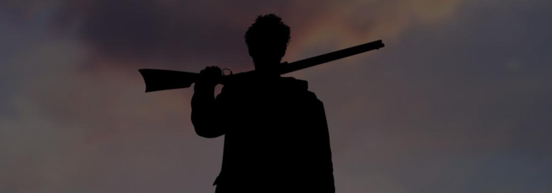 photo of silhouette of a man standing with his legs apart holding a rifle across his shoulders against a cloudy horison